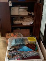 Wide view showing wooden cabinet with shelves of folded white linens plus placemats leaning inside cabinet and fabric pieces in plastic bin.