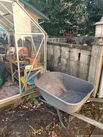 Metal wheelbarrow next to greenhouse showing overall condition, some dirt and leaves inside.