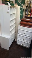 Photo showing white storage pieces including a tall shoe organizer with compartments and a three-drawer nightstand next to a smaller white storage unit.