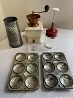 Two metal baking trays with shell-shaped molds, a metal grater, a wooden coffee mill, and a manual jar chopper on table