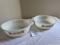 Two Fire-King casseroles shown side by side on a white cloth. Both have the Harvest wheat pattern in yellow and brown with white milk glass.