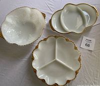 Photo showing three Fire-King milk glass serving dishes: oval three-section divided tray with gold rim, round three-section divided dish with gold and beaded rim, and scalloped grape salad bowl with embossed grape design and gold rim.