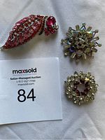 Three assorted brooches arranged on fabric with auction lot card: pink rhinestone leaf-shaped brooch, aurora borealis starburst brooch, clear rhinestone wreath-shaped brooch.