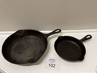 Two cast iron skillets side by side on a white surface; one larger and one smaller, with handles and pouring spouts.