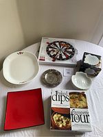 Photo showing the entire lot on table including Stokes dartboard cheese platter set, Gourmet Village baked dips box, 8 coquilles St-Jacques shells in packaging, white shell dish, red square dish, Corelle oval platter, WmA Rogers wine bottle holder and silver plate.