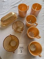 Photo showing 3 peach lustre mugs and 4 amber glass pieces including a butter dish, creamer and sugar bowl.