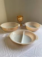 Four Fire-King glass items photographed on neutral background including one round casserole, one oval casserole, one divided oval casserole, and one amber goblet