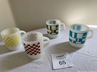 Four Fire-King milk glass mugs with different colored geometric and checkered designs sitting on a white cloth.