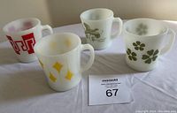 Four Fire-King opaque white glass mugs with different colorful designs including floral, geometric, and bird motifs on a table