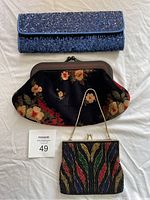 Three handbags laid out flat on a fabric background: a blue sequined rectangular clutch, a black floral Estro di Firenze clutch, and a beaded GeoMercier Montral clutch with multi-color pattern and chain handle.