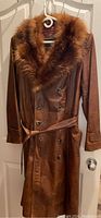 Full view of the vintage brown leather coat with fur collar hanging on a hanger, displaying front and length.