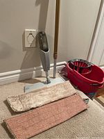 Norwex mop with adjustable handle and spray bottle, two mop cloth pads (beige and rust-colored), and red Vileda Quick Wring bucket photographed on carpet.