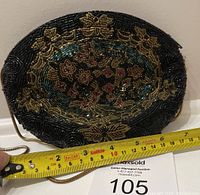 Front view of oval black beaded purse with floral beadwork and gold-tone metal chain strap partially visible as measured with tape showing approx. 7 inches wide