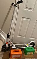 White Gymax scooter with adjustable handlebar and black deck leaning against door with two rectangular snow brick makers on floor in front