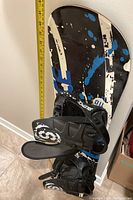 Side view of the snowboard showing the graphic design and one binding attached. A measuring tape indicates the length of the snowboard is slightly under 60 inches.