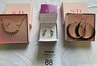Three Stella & Dot jewelry items in their original boxes: gold-tone necklace with spikes and crystals, small gold and crystal reversible drop huggie earrings, and large gold-tone feather detail hoop earrings.