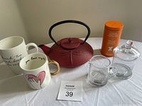 Photo showing all items in the lot including red cast iron tea pot with black handle, two ceramic Indigo brand mugs, orange tea tin, clear glass creamer, and clear sugar bowl.