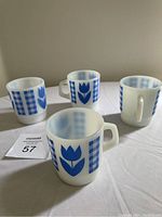 Four white opaque glass mugs each with a blue tulip and gingham pattern on the sides and square handles.