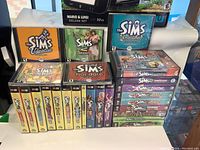 Front image showing a collection of Sims PC game cases including main games and expansion packs neatly stacked, with some discs visible inside jewel cases.