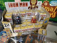 Photo of majority of 26 Iron Man Micro Muggs figures in retail box packaging showing front and top rows with two visible figures standing above boxes: one black and red armored figure and one classic red and gold Iron Man figure.