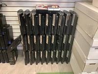 Multiple Sony PS3 fat model consoles arranged upright on a floor against a wall, showing front panels with disc trays and slots. Some consoles appear dusty and worn.