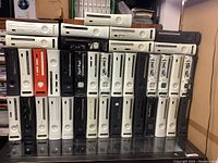 Front view of approximately 37 Xbox 360 consoles stacked in rows with various colors visible including white, black, and orange. Many consoles show different states of wear including missing or damaged disc trays.