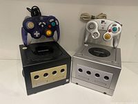 Two Nintendo GameCube consoles side by side with two controllers placed on top (one indigo, one silver).