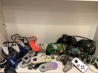 Lot of various gaming controllers shown together on shelf with cables tangled, various makes, colors and styles visible.