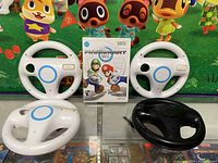 Mario Kart Wii game case centered behind four Wii steering wheel controllers (three white, one black) on green Animal Crossing backdrop.