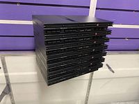 Photo showing a stack of eight black Sony Playstation 2 SCPH-70011 consoles with visible front ports and ventilation.