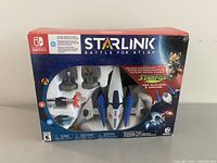 Front of the box showing Starlink Battle for Atlas starter pack with detailed Arwing spaceship toy model, Fox McCloud figure, and accessory weapons visible inside packaging.