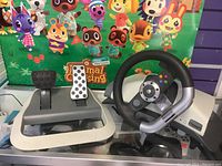 Front view of Xbox 360 racing wheel and pedals on glass surface with Animal Crossing poster background.