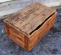 Closed view of a large wooden crate with a lid that has stenciled text reading 'HANDLE WITH CARE' and other faded labels. The crate is rectangular and shows signs of weathering and use.