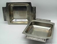 Two square stainless steel serving and baking dishes with decorative side handles. Both dishes show the exterior and interior surfaces with a brushed finish and embossed accents on handles.