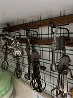 Photo showing multiple antique hand beaters hanging on wooden rack against a checkered background, displaying different sizes and metal gear mechanisms.