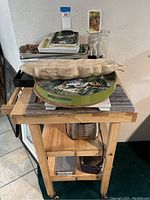 Wooden butcher block cart with kitchen items stacked on top and on shelves, including trays, cutting boards, glass jar, and toaster.
