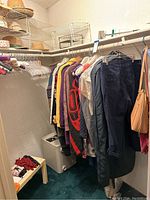 Clothing hanging on plastic and cloth-covered hangers with sweaters, outerwear, pants visible. White cubed safe below clothes.