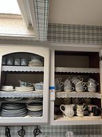 Open kitchen cabinet showing neatly arranged blue and white teacups, saucers, dinner plates, salad plates, bowls, several ceramic mugs, cream pitcher, and other kitchenware items on lace-trimmed shelves.