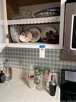 Kitchen cabinet with shelves holding ceramic plates, bowls, and decorative items above countertop with assorted glass and metal barware.