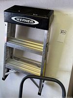 Werner brand aluminum step ladder with black top, two wide steps, and safety braces connected to the frame.