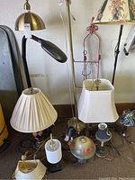 Photo showing multiple lamps of different styles and sizes including brass dome-shaped lamp, black adjustable desk lamp, tall floor lamp with floral shade, and others.