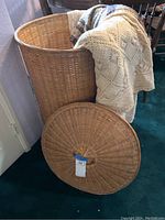 Large woven basket with lid removed, sitting on floor with cream embroidered tablecloth draped over basket, green carpet and wooden table in background.