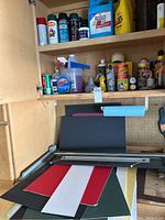 Spray paint and auto care products on shelves above a matte cutter and various colored matting boards lying on a work surface in a garage.
