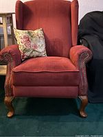 Front view of burgundy upholstered reclining chair with floral pillow on seat, showing hand-nailed metal tacks on armrests and wooden Queen Anne feet.