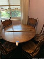 Round oak dining table with five matching chairs placed around it near a window with curtains.