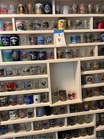 Wide view of multiple shelves filled with various shot glasses, showing different city names, logos, and colorful images printed on them.
