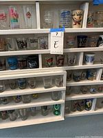 Shot glasses on shelves showing detailed logos, landmarks, cities, and themes, including Amtrak, Notre Dame, and various scenic designs.