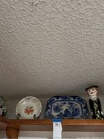 Wide view showing a wooden shelf with the decorative ceramic pieces placed on top: large blue and white rectangular platter center back, floral plate to the left, ceramic figure to the right.