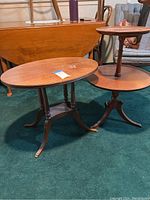Photo of two antique wooden tables on a green carpet; one oval Maple table and one two-tiered table with pie crust edging.