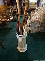 Overall view of the white ceramic holder containing four umbrellas and three walking sticks in an indoor setting with furniture.